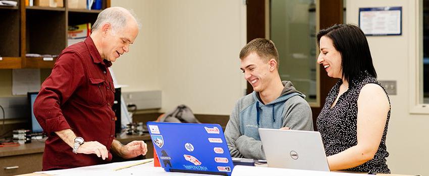Two professors smiling with student in classroom looking at laptop